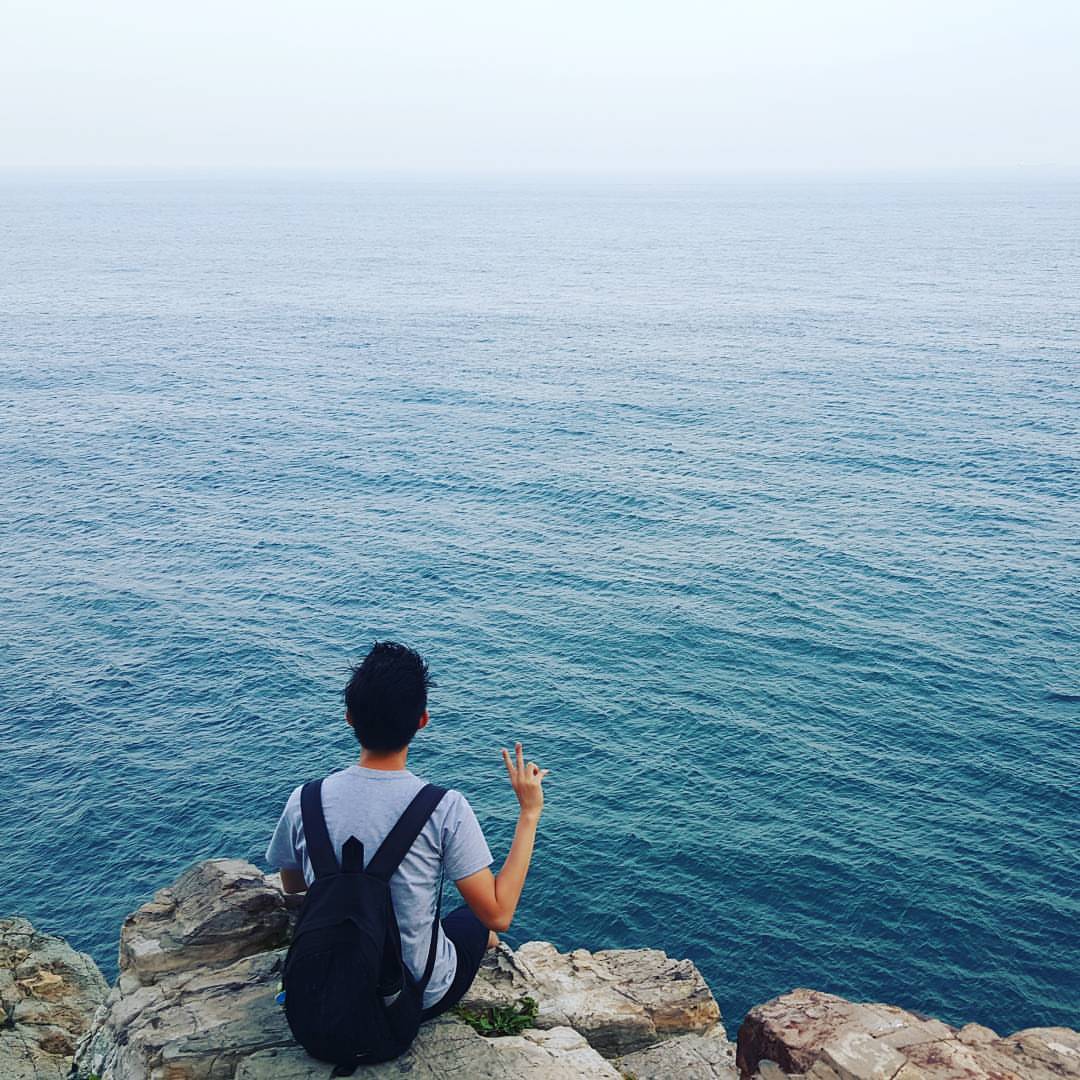 A photo from behind Endi's back, with Endi sitting on a rock cliff, facing the vast sea. His right hand forms a V pose.